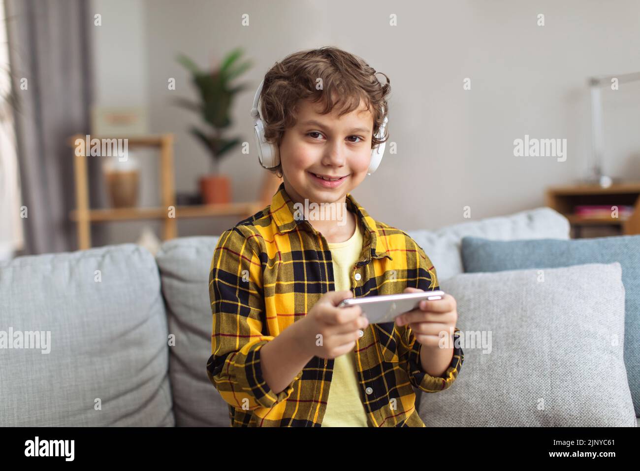Piccolo giocatore. Ritratto in interni di un simpatico ragazzo che indossa cuffie per giocare a videogiochi sullo smartphone a casa Foto Stock