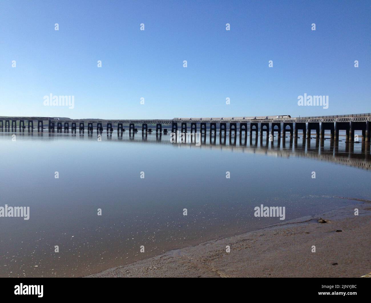 Un treno sulla costa orientale che attraversa il Tay Bridge, con riflessione in acqua in condizioni calme sotto il cielo blu a Dundee, Scozia, Regno Unito. Foto Stock