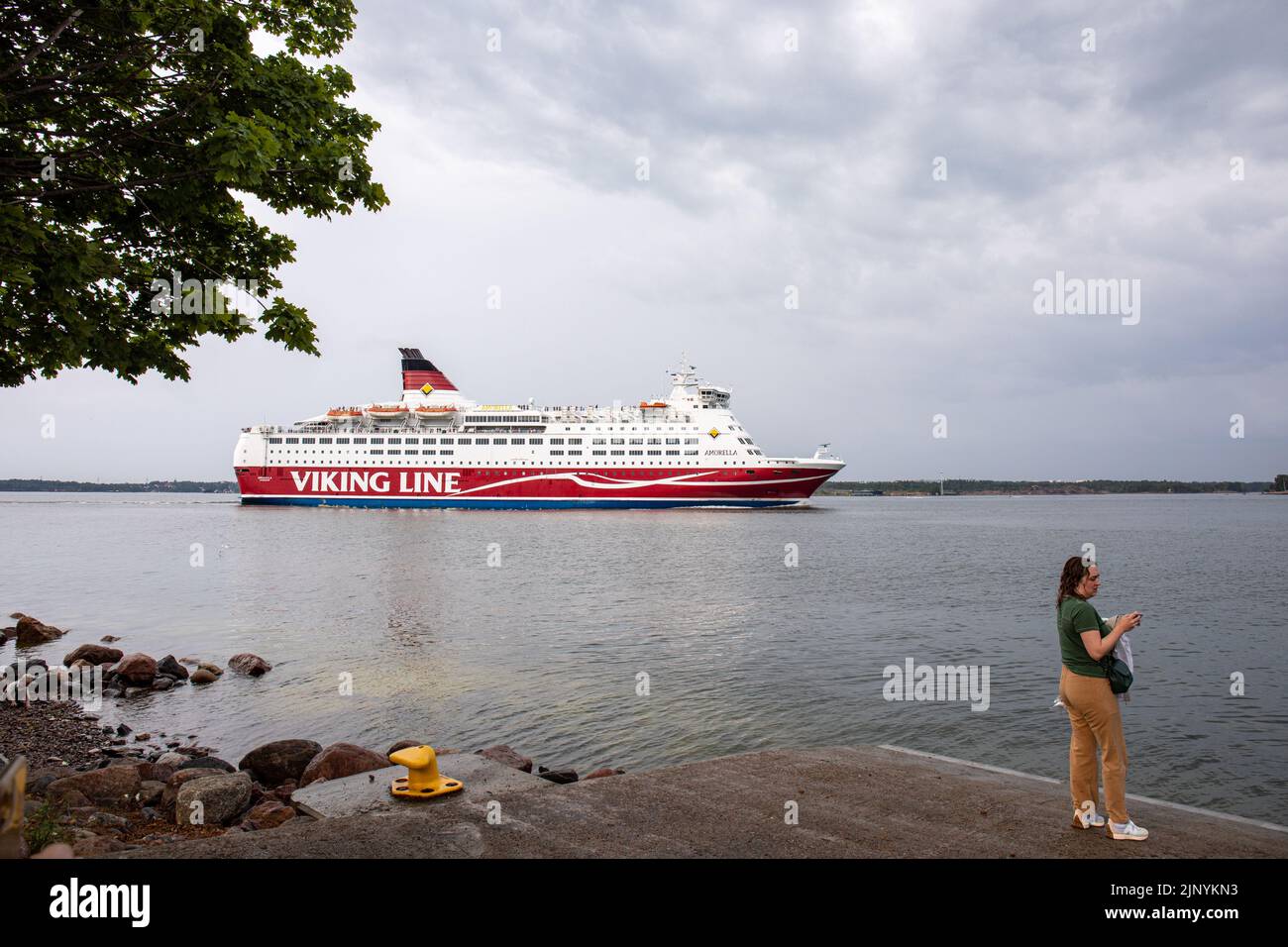 Donna in piedi sul molo mentre il traghetto da crociera M/S Amorella della compagnia di navigazione Viking Line passa l'isola di Lonna a Helsinki, Finlandia Foto Stock
