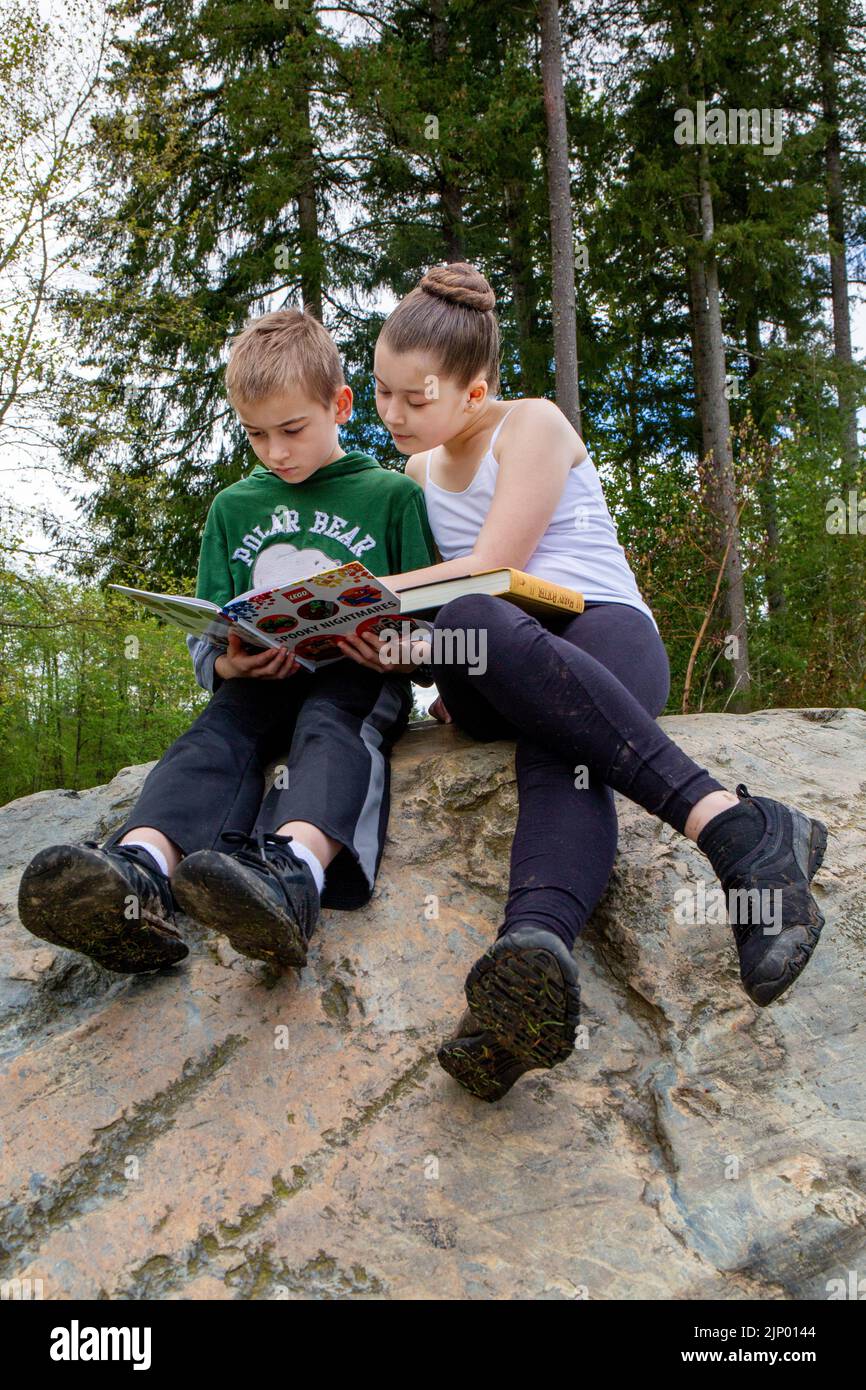 Issaquah, Washington, Stati Uniti. sorella di 11 anni e suo fratello di 9 anni che legge, con la sorella che lo aiuta, seduto in cima a un grande masso in un par Foto Stock
