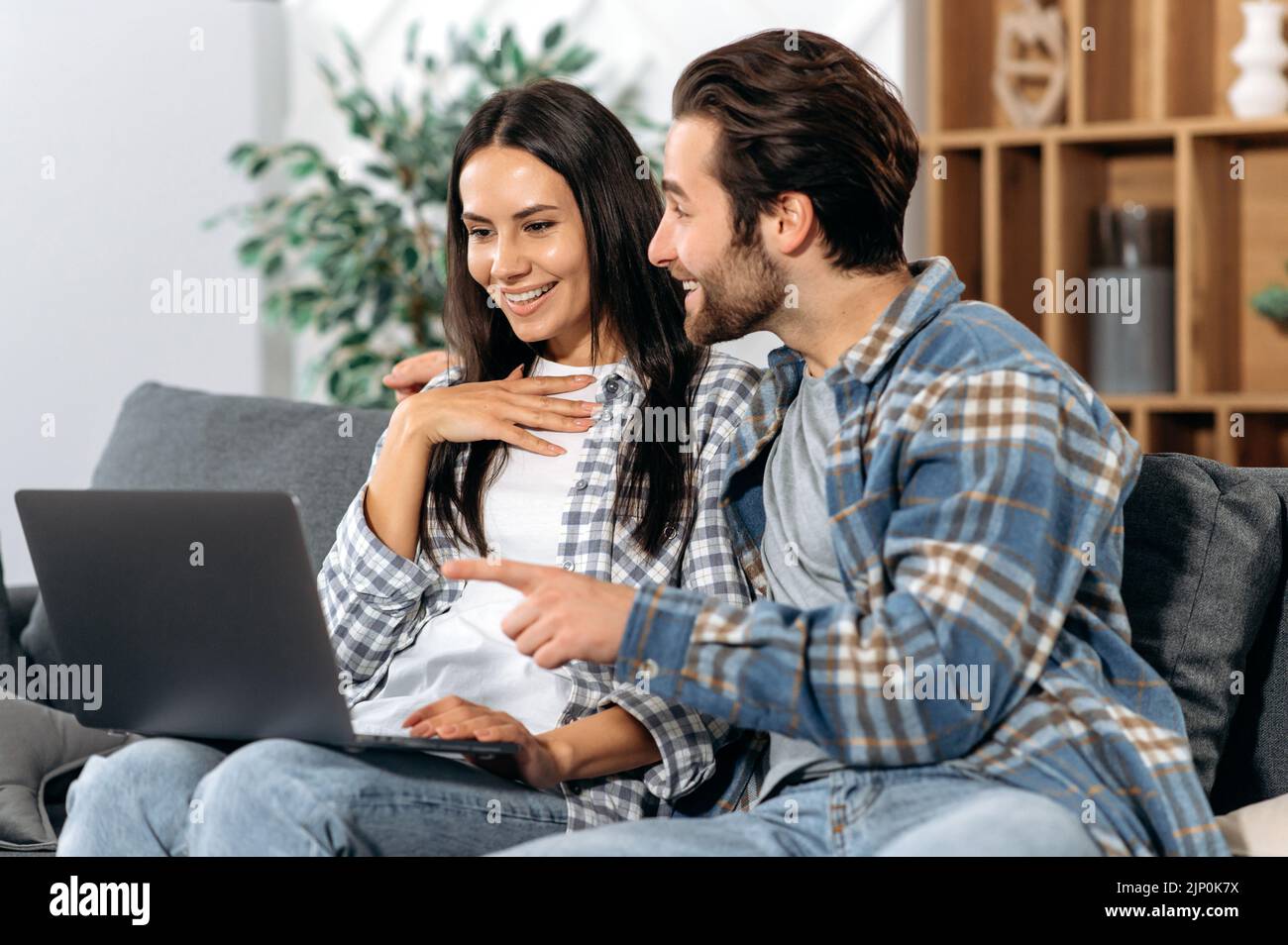 Sposi positivi che trascorrono del tempo insieme a casa, seduti sul comodo divano del soggiorno, utilizzano un notebook, effettuano acquisti online, guardano le notizie, sfogliano i social network, sorridi insieme Foto Stock