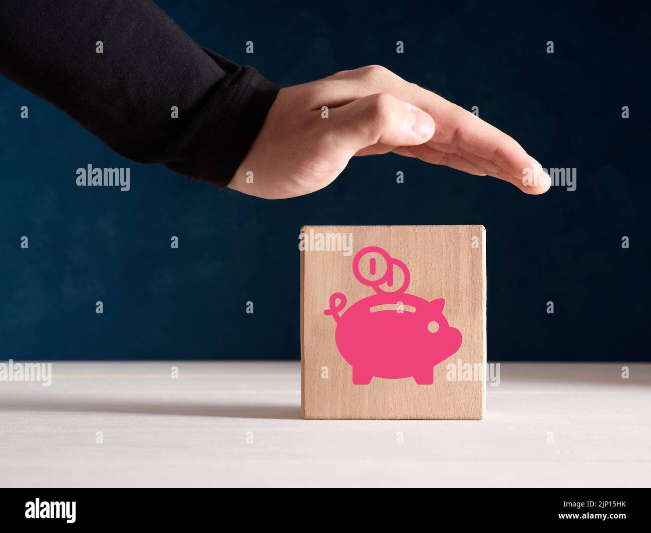 Pianificazione finanziaria, risparmio di denaro, assicurazione e concetto di protezione. La mano maschile si erge in modo protettivo sopra il blocco di legno con l'icona di un piggy bank. Foto Stock