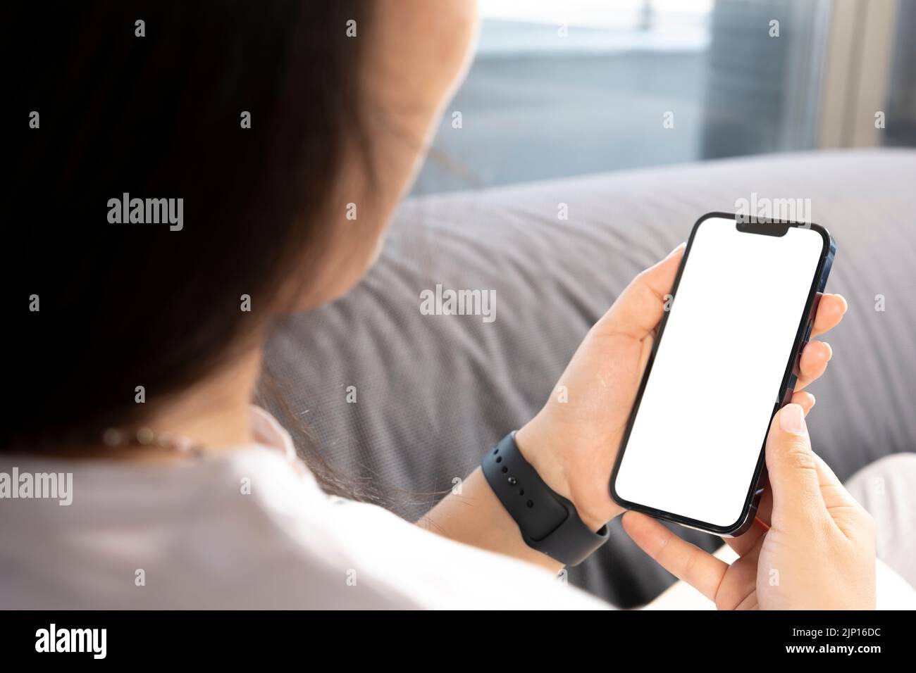 Con uno smartphone, una giovane donna seduta sul divano e uno smartphone con un display bianco mockup vuoto. Schermo vuoto verticale per le app dei social media. Foto Stock