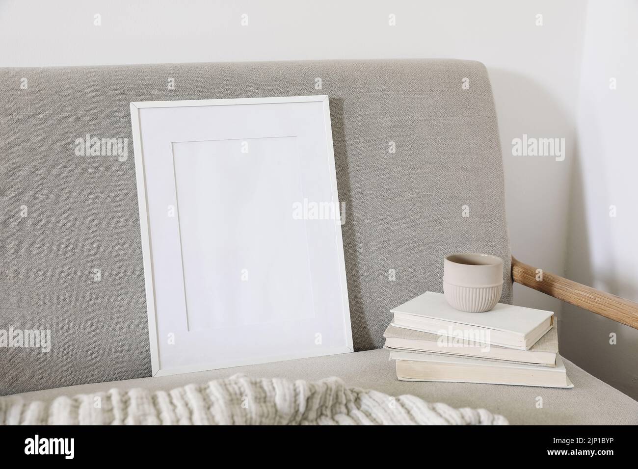 Cornice verticale vuota in legno mockup. Tazza di caffè su un mucchio di vecchi libri. Divano in lino di metà secolo. Sfondo bianco parete. Concetto di home Office Foto Stock