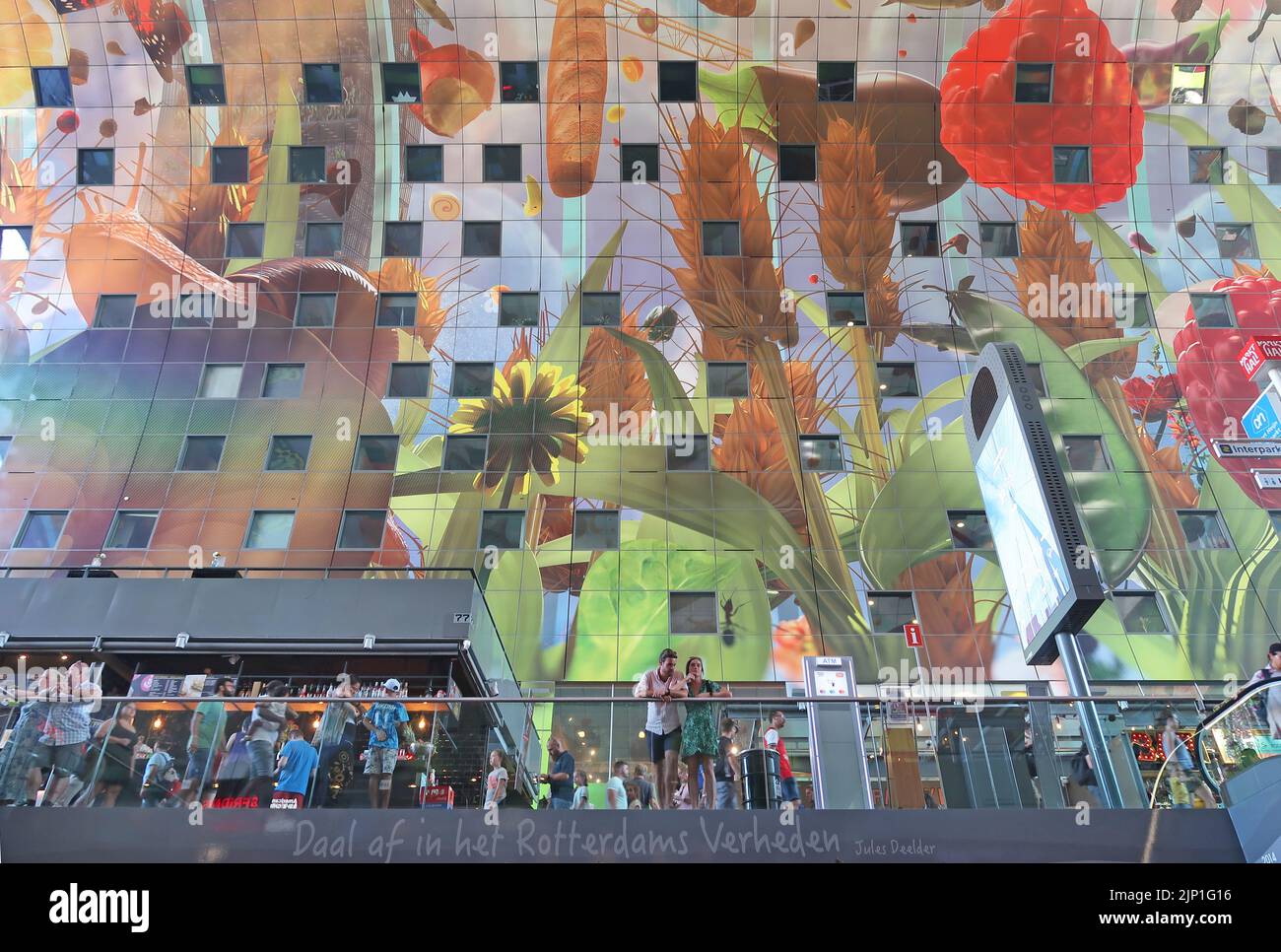 La vista interna del Rotterdam Markthall (mercato coperto) mostra il soffitto curvo con il suo gigantesco murale con frutta e verdura. Coppia in primo piano. Foto Stock
