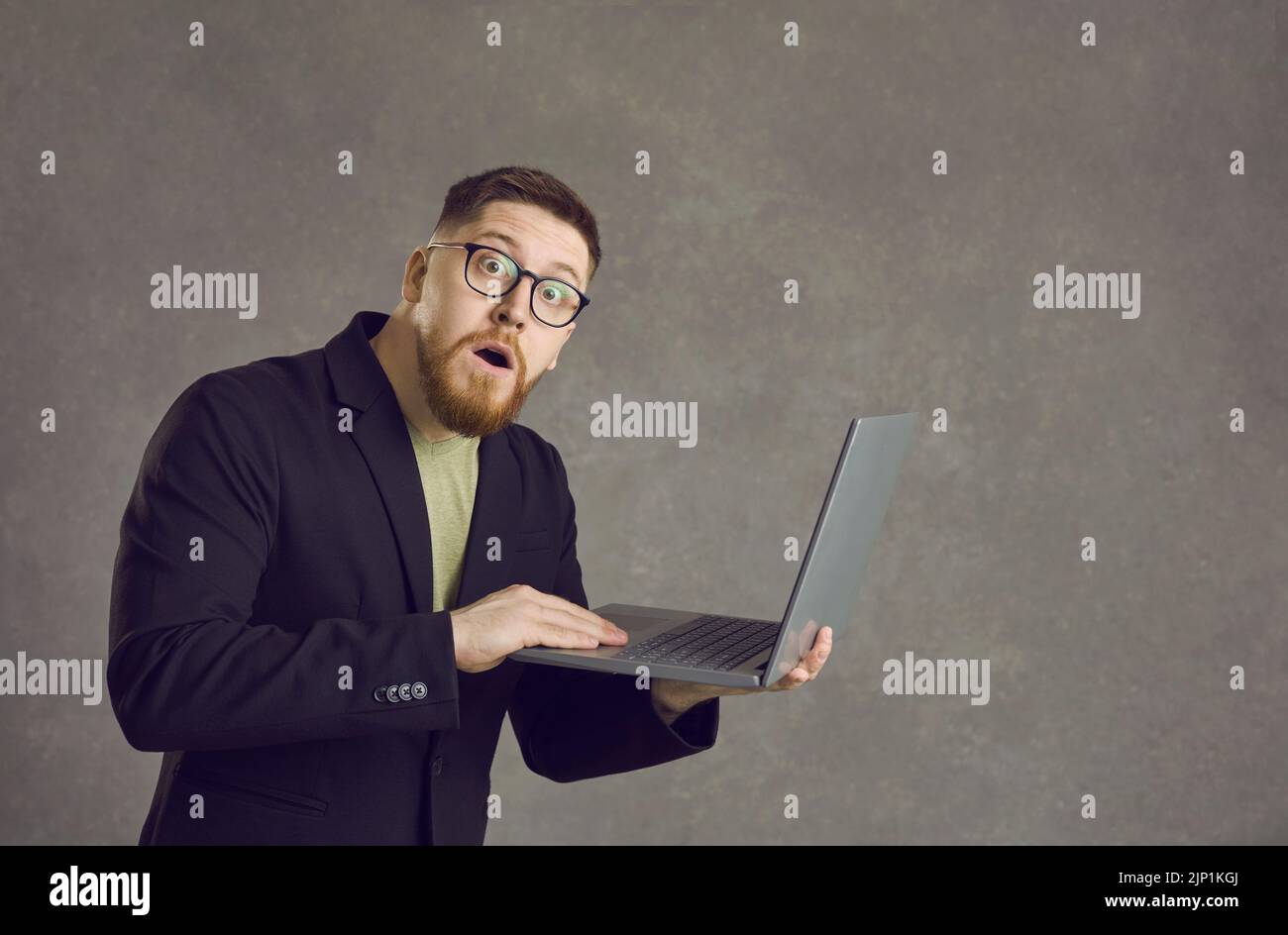 Ritratto di un barbuto scioccato uomo in piedi con un computer portatile in mano su uno sfondo grigio. Foto Stock