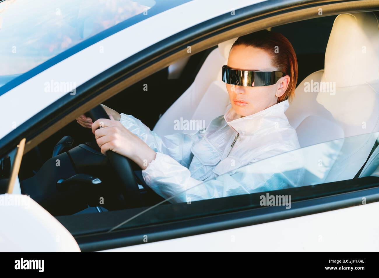 Ritratto di moda cyberpunk ragazza alla guida di una supercar bianco. Donna capelli rossi con occhiali futuristici si siede accanto al volante dell'auto. Futuro tecnolo Foto Stock