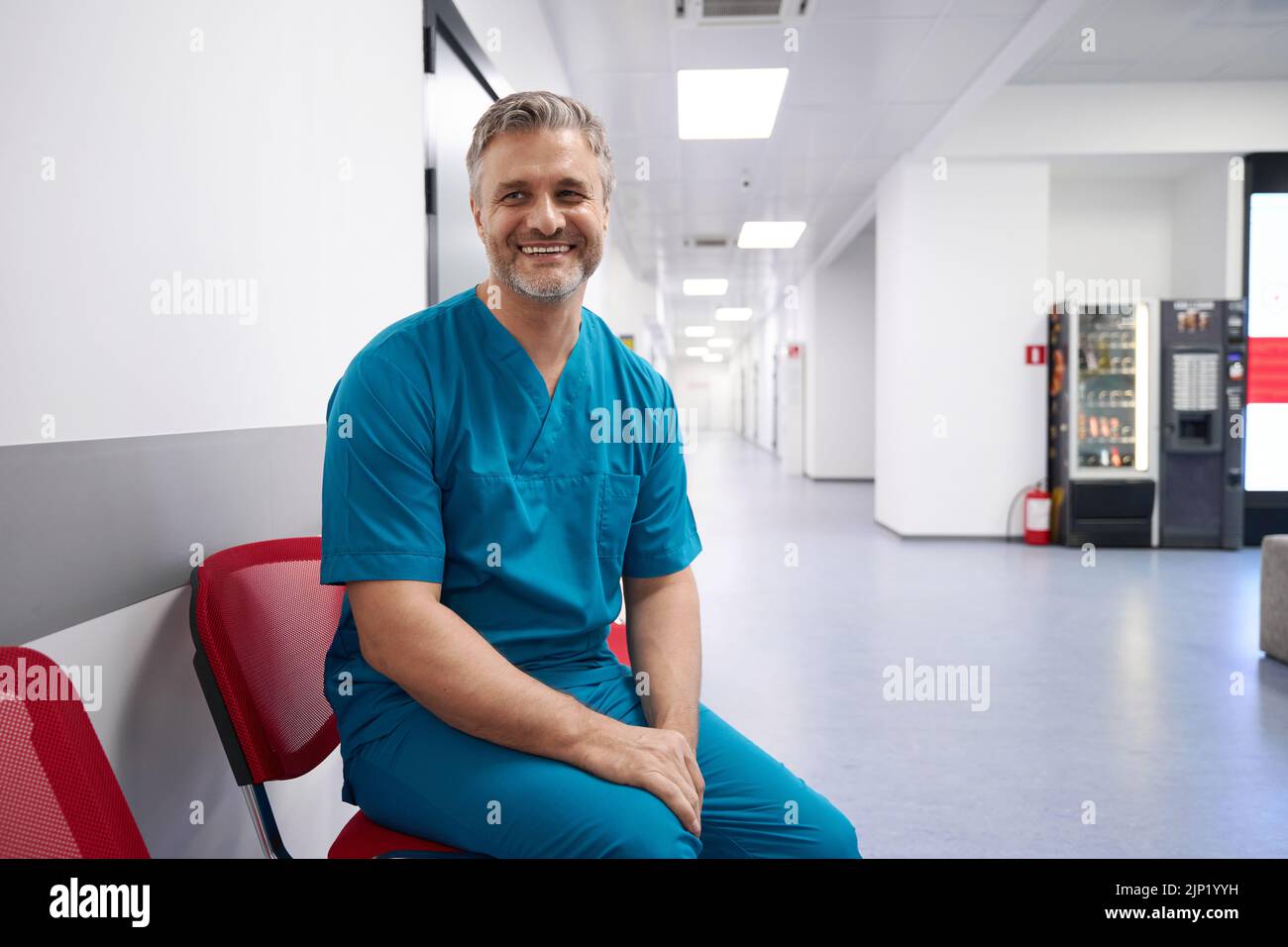 Dottore in uniforme blu sorridente mentre si siede nella lobby dell'ospedale Foto Stock