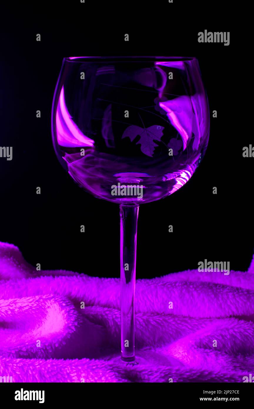 Bicchiere di vino vuoto su una morbida coperta con retroilluminazione viola. Composizione astratta. Concetto di cocktail alcolico. Foto Stock