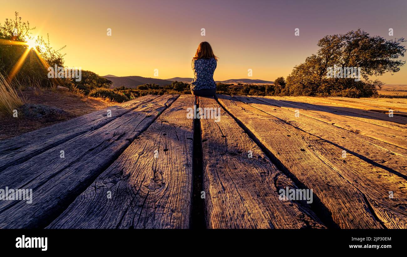 Donna seduta sulla schiena su pavimento di legno all'aperto e godersi il tramonto sul campo. Spagna. Foto Stock