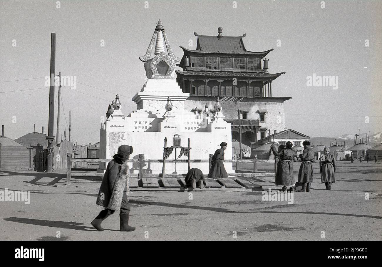 1960s, storico, molti mongolesi maschi in piedi - uno starnutendo giù su una piattaforma di legno - ad uno stupa al Monastero di Gandan a Ulaanbaatar, Mongolia, Asia Centrale. Lo stupa è un momumento sacro per la mediazione e la preghiera. Il vecchio tempio buddista, il tempio di Janraiseg, può essere visto dietro il santuario. Il Monastero fu fondato nel 1809, quando una scuola per lo studio del Buddismo superiore si separò dal Monastero di Bogd Gegenov e fu chiamato Gandan, in onore di Gaden, un monastero buddista tibetano. Foto Stock
