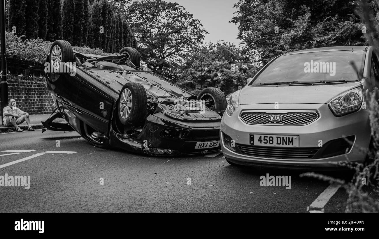 Un'immagine in bianco e nero di un conducente e della sua auto ribaltata su una strada a Londra. Foto Stock
