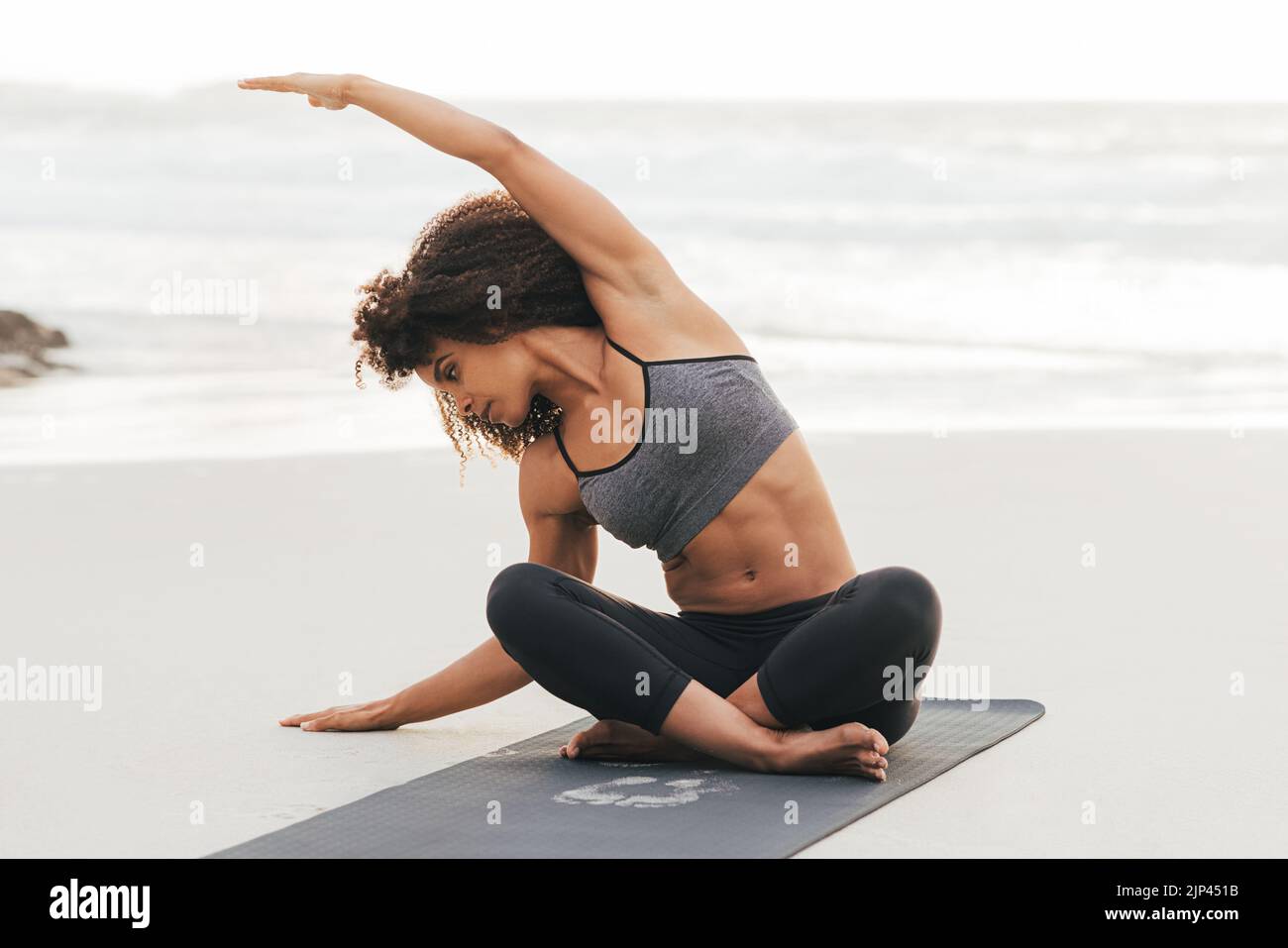 Donna in abbigliamento sportivo che fa esercizi di flessibilità mentre si siede su un tappetino su una spiaggia Foto Stock
