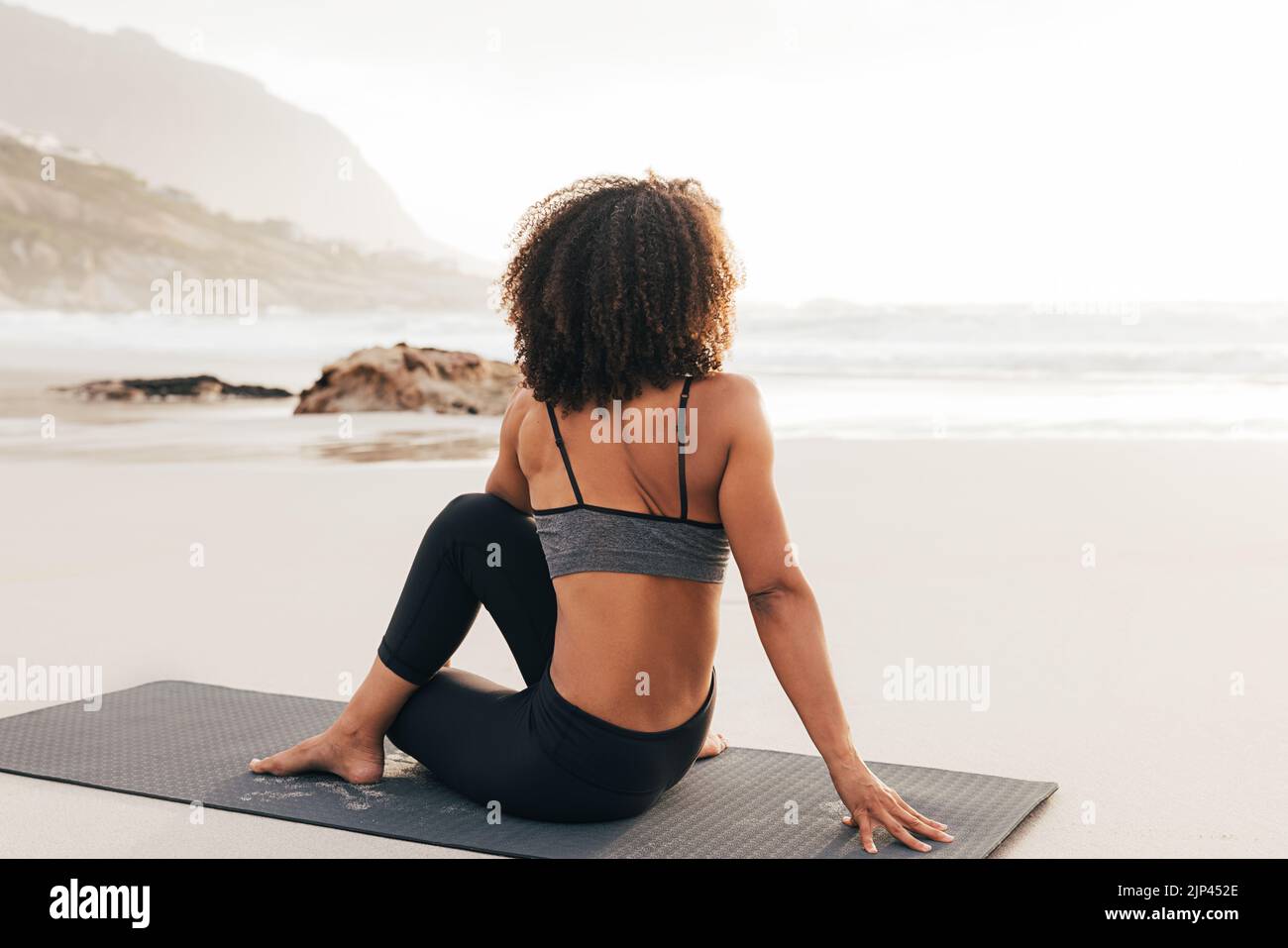 Donna seduta su un tappetino sulla spiaggia al tramonto. Vista posteriore di una donna che si rilassa dopo gli esercizi di yoga. Foto Stock