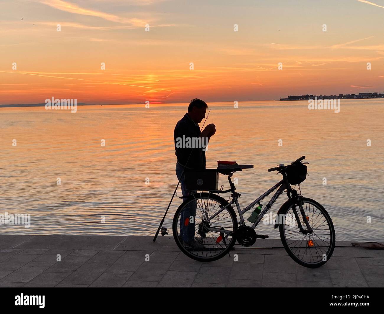 Una silhouette di maschio con una canna da pesca e una bicicletta sulla costa al tramonto Foto Stock