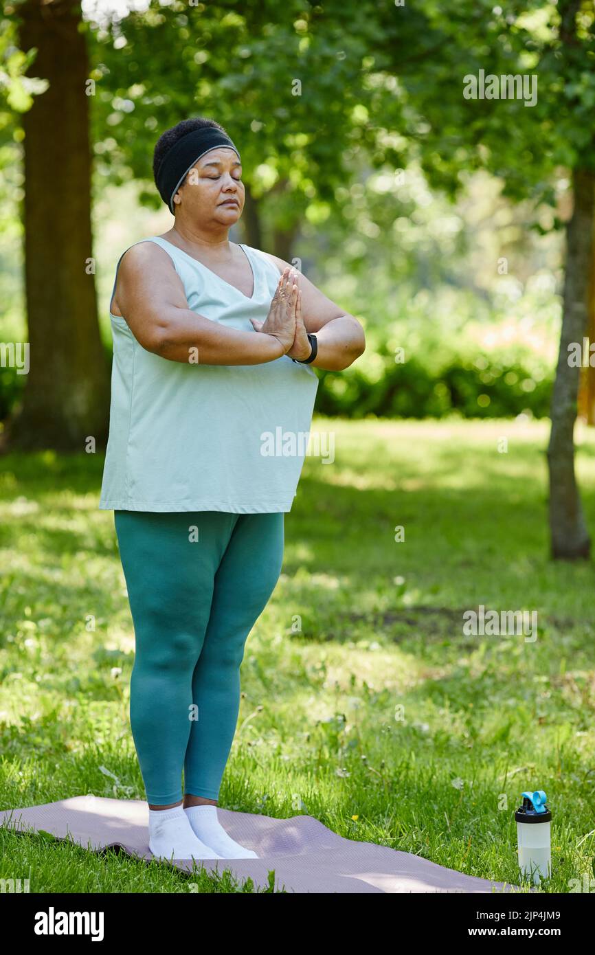 Verticale ritratto completo di donna nera matura godendo yoga all'aperto nel parco verde e in piedi con gli occhi chiusi facendo esercizi di respirazione Foto Stock