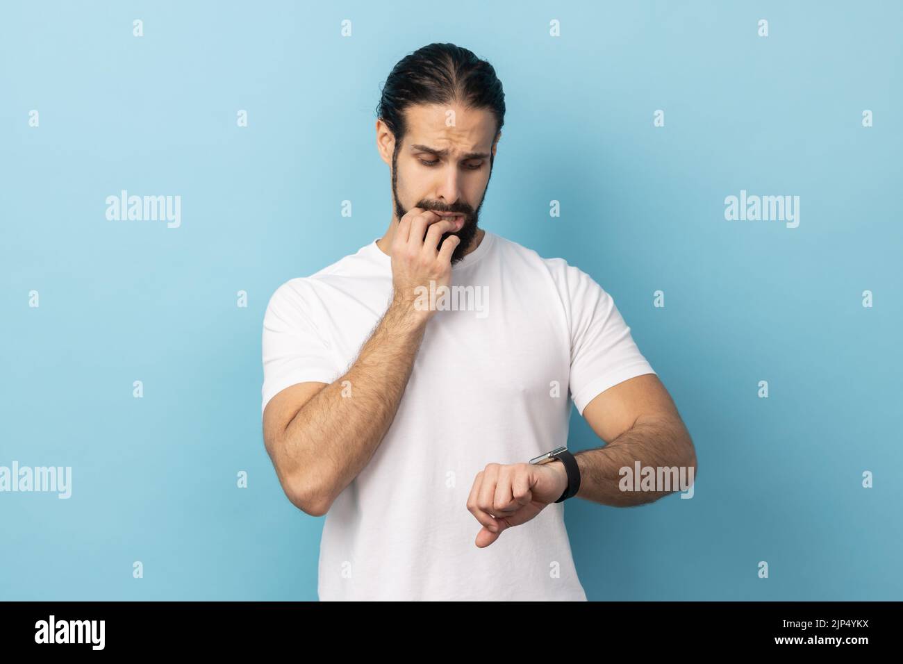 Ritratto dell'uomo con la barba che indossa una T-shirt bianca che punta il dito sul suo orologio da polso con il viso nervoso, il tempo è fuori, mordente unghie. Studio in interni isolato su sfondo blu. Foto Stock