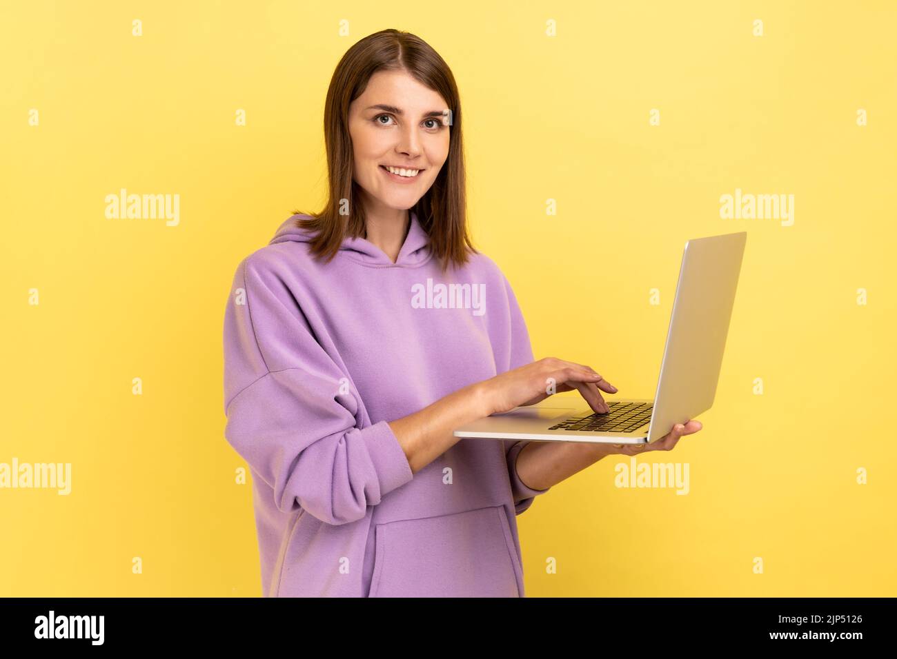 Ritratto di una giovane adulta ottimista e positiva che lavora al computer portatile, guardando la fotocamera con un volto felice, con felpa con cappuccio viola. Studio al coperto isolato su sfondo giallo. Foto Stock