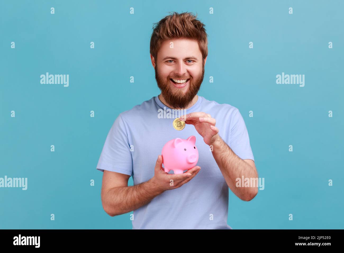 Ritratto di felice uomo sorridente bearded guardando la macchina fotografica, mettendo moneta in piggybank, risparmio di denaro, esprimere emozioni positive. Studio in interni isolato su sfondo blu. Foto Stock