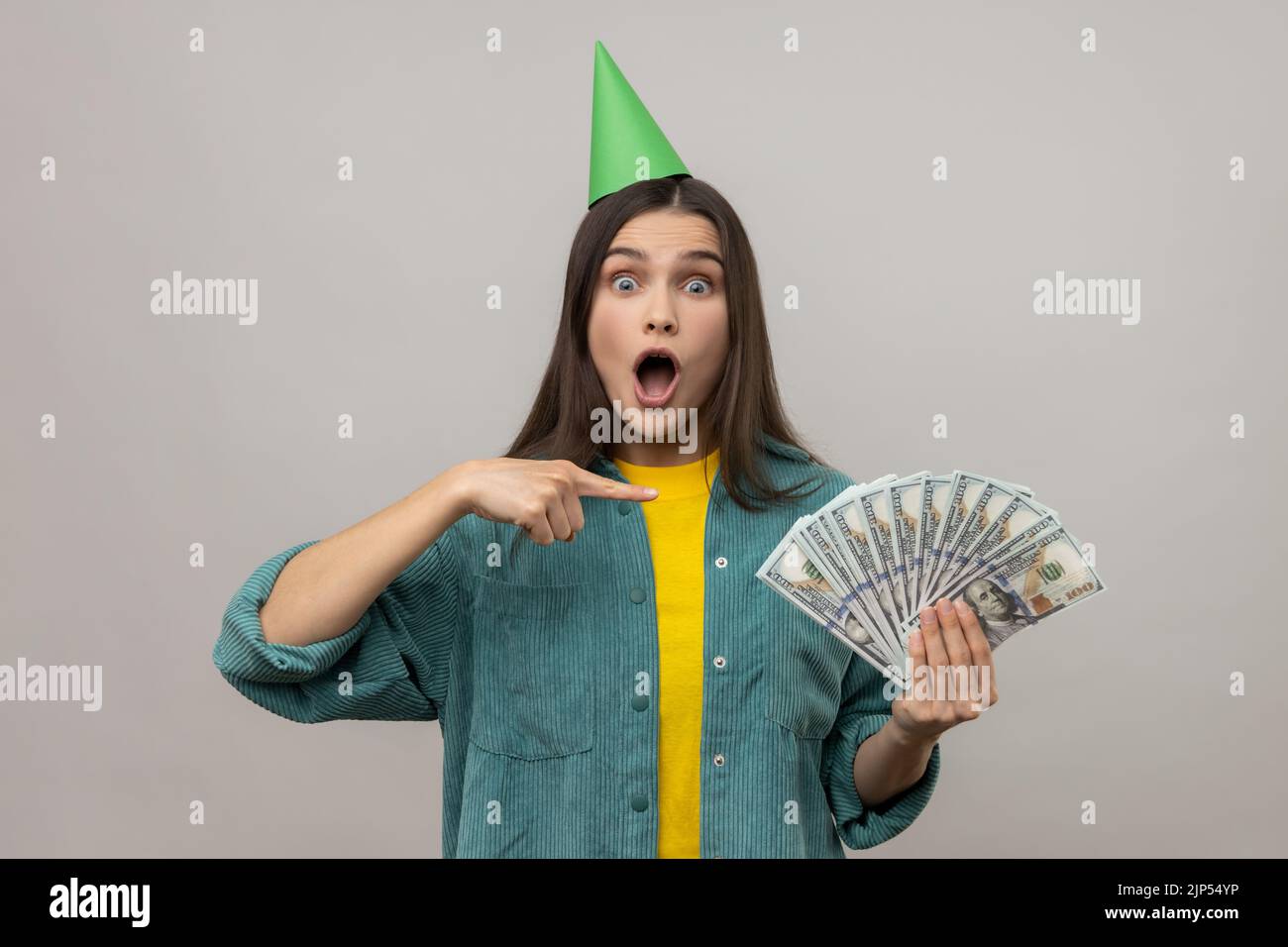 Stupito eccitato ricca donna in festa cono sulla testa che punta i soldi, osservando sorpresi dalla vittoria della lotteria, celebrando il profitto grande, portando la giacca casual di stile. Studio in interni isolato su sfondo grigio Foto Stock