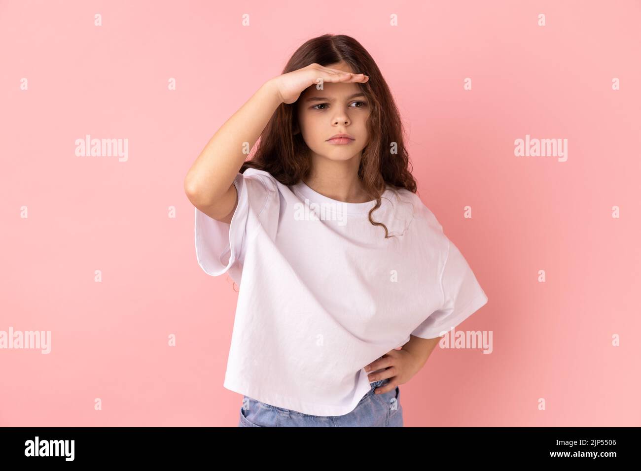 Ritratto di bambina che indossa una T-shirt bianca che sbircia in lontananza, guardando lontano con una vista attenta, cercando all'orizzonte e curiosi di scoprire. Studio in interni isolato su sfondo rosa. Foto Stock