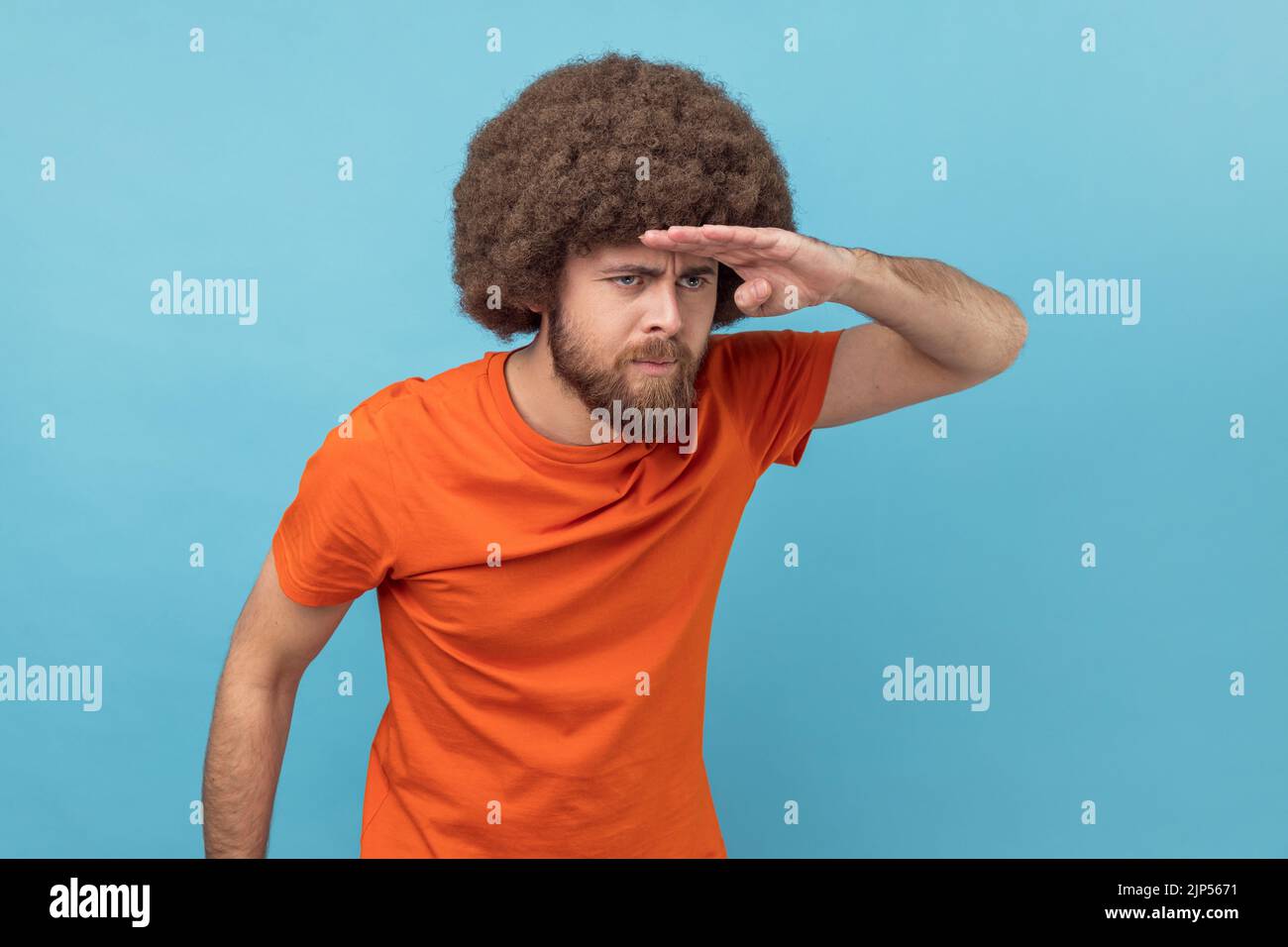 Ritratto dell'uomo con acconciatura afro che indossa una T-shirt arancione che guarda lontano con la mano sopra la testa, cercando con attenzione un futuro luminoso. Studio in interni isolato su sfondo blu. Foto Stock