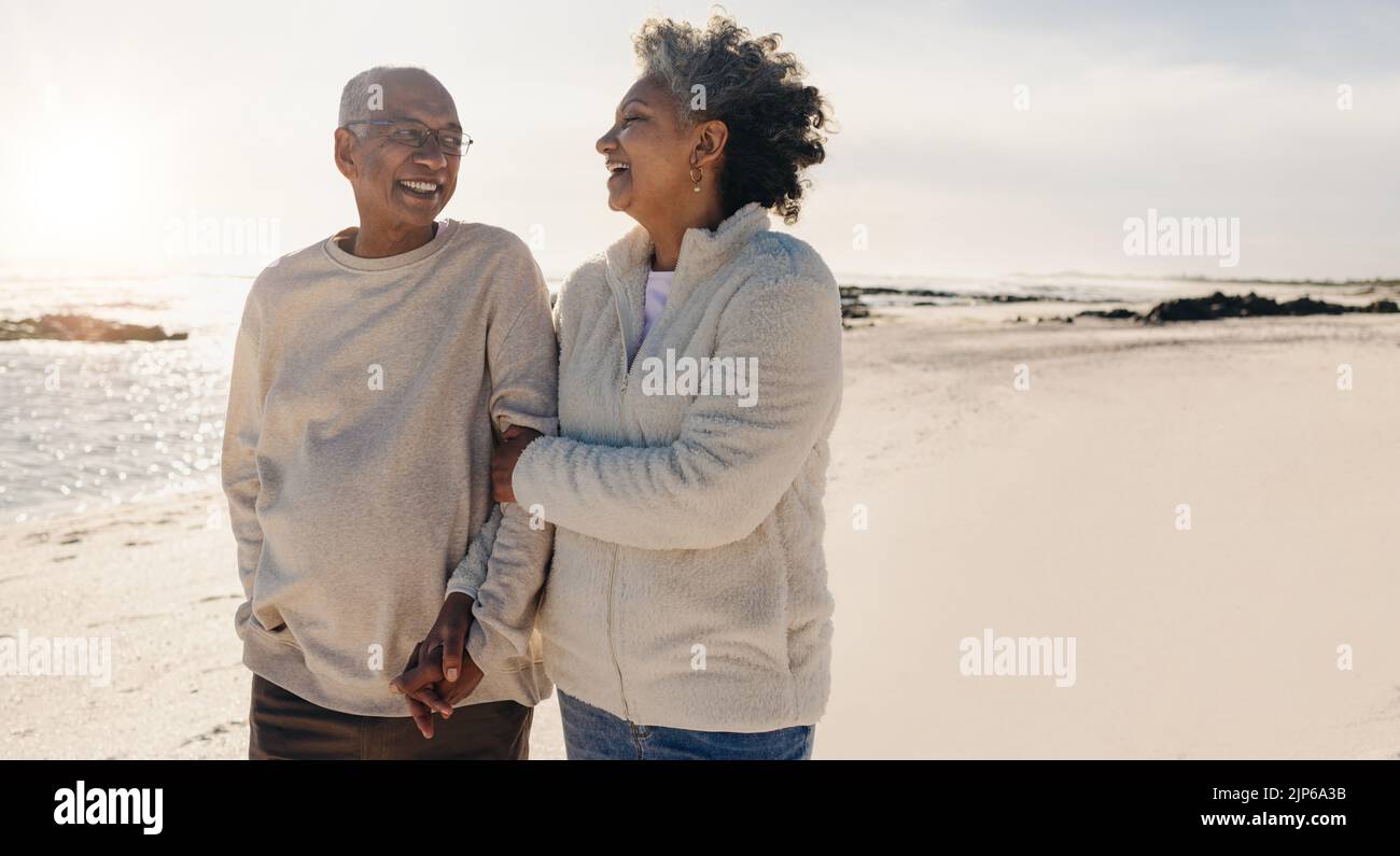 Romantica coppia anziana che ride insieme mentre si fa una rinfrescante passeggiata lungo la spiaggia. Coppia anziana felice godendo i loro giorni di pensione a. Foto Stock
