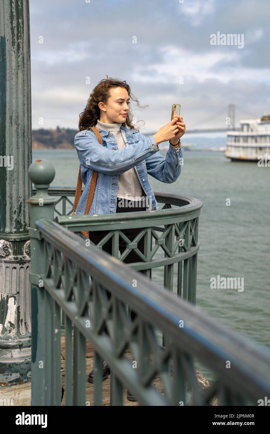 Young Woman on Pier con vista sulla baia di San Francisco per scattare foto con lo smartphone Foto Stock
