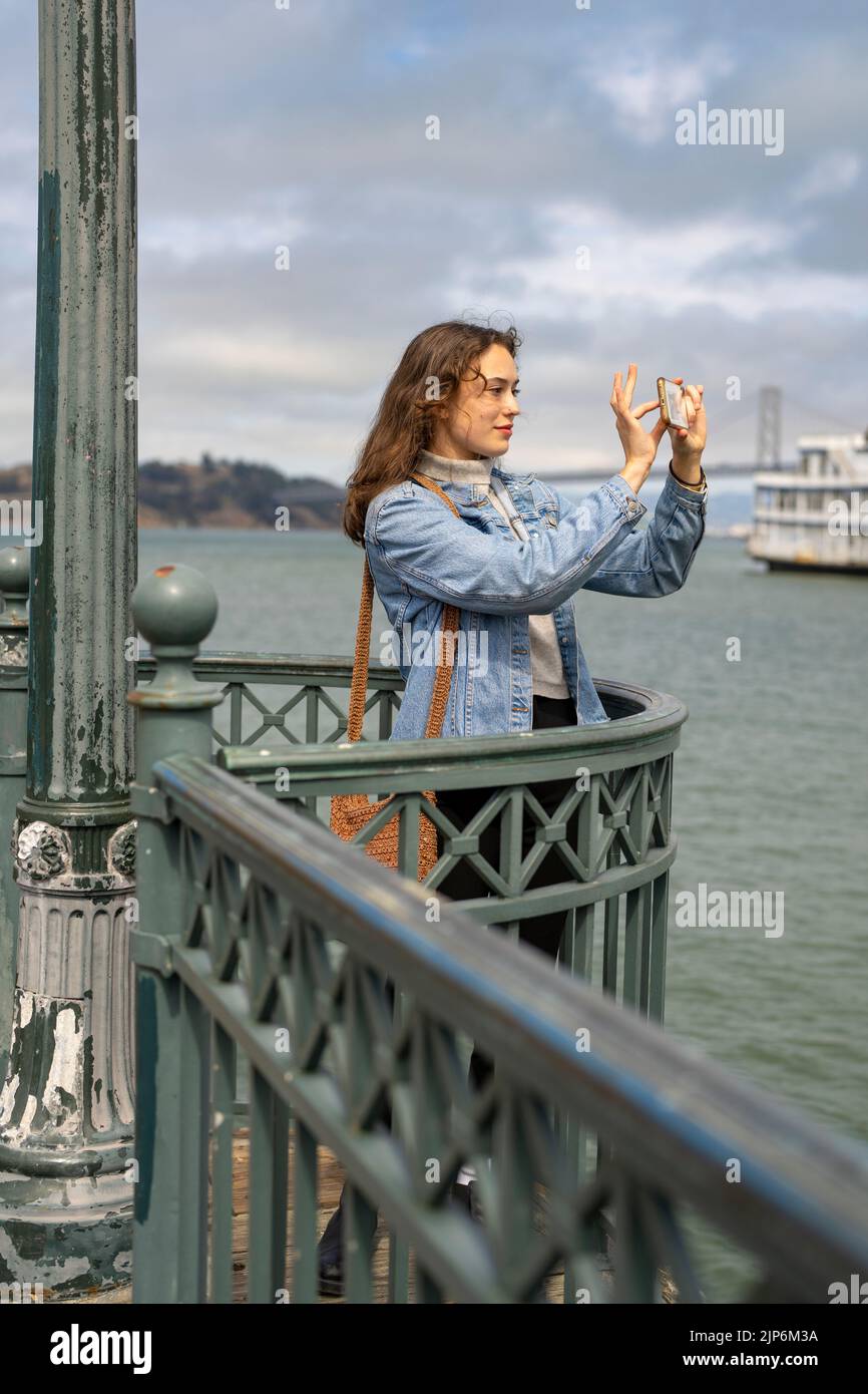 Young Woman on Pier con vista sulla baia di San Francisco per scattare foto con lo smartphone Foto Stock