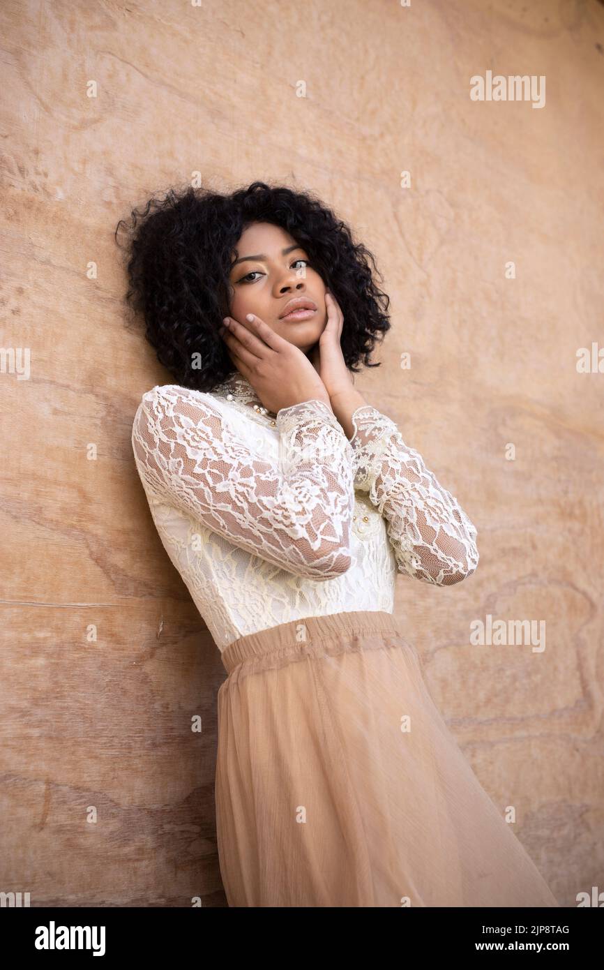 Dal basso di aggraziata giovane afro-americana modello femminile con capelli ricci scuri in romantico vestito toccare guance e guardare la fotocamera mentre si appoggia Foto Stock