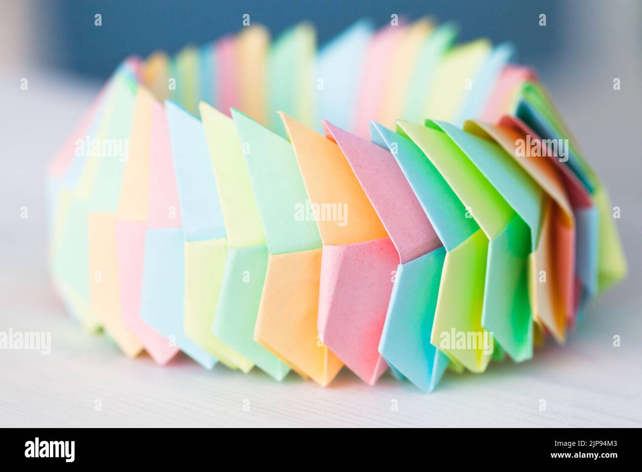 L'anello Origami in fogli di carta colorati collegati giace su un tavolo di legno bianco, foto ravvicinata con messa a fuoco morbida selettiva Foto Stock