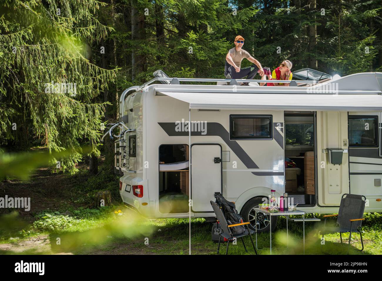 Famiglia di due, marito e moglie, che riposano sul tetto del loro Camper White Van parcheggiato nel mezzo della Scenic Green Forest godendo il momento durante Foto Stock