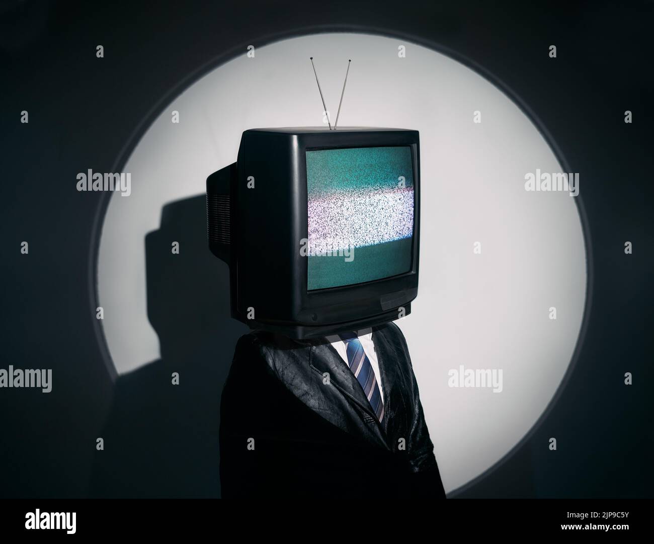 Uomo con la TV invece della testa. Media zombie concetto con televisione addicted maschio in nero cappotto con braccia incrociate in posa in studio sotto i riflettori. Con Foto Stock