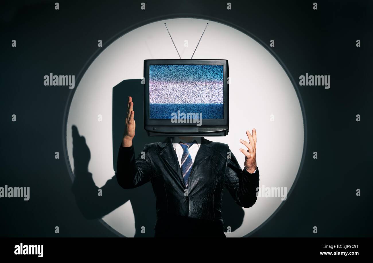 Uomo con la TV invece della testa. Media zombie concetto con televisione addicted maschio in nero cappotto con braccia incrociate in posa in studio sotto i riflettori. Con Foto Stock