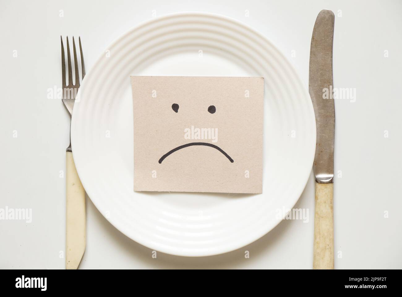 Un emoticon triste si trova in un piatto su uno sfondo bianco, e accanto ad esso è un coltello con una forchetta, un cattivo umore, mancanza di cibo, fame e dieta Foto Stock