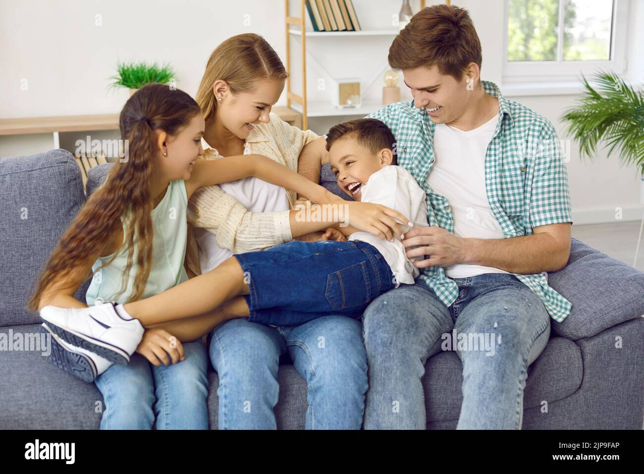Genitori e bambini felici si solleticano mentre si siedono insieme sul divano a casa. Foto Stock