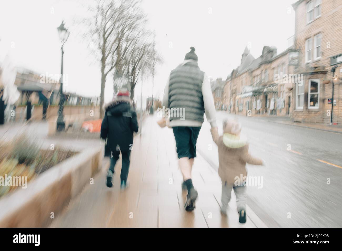 Uomo che indossa pantaloncini corti con bambini che camminano per strada in una giornata invernale bagnata, tenendo le mani. Centro di Ilkley, West Yorkshire, Inghilterra, Regno Unito (ICM) Foto Stock