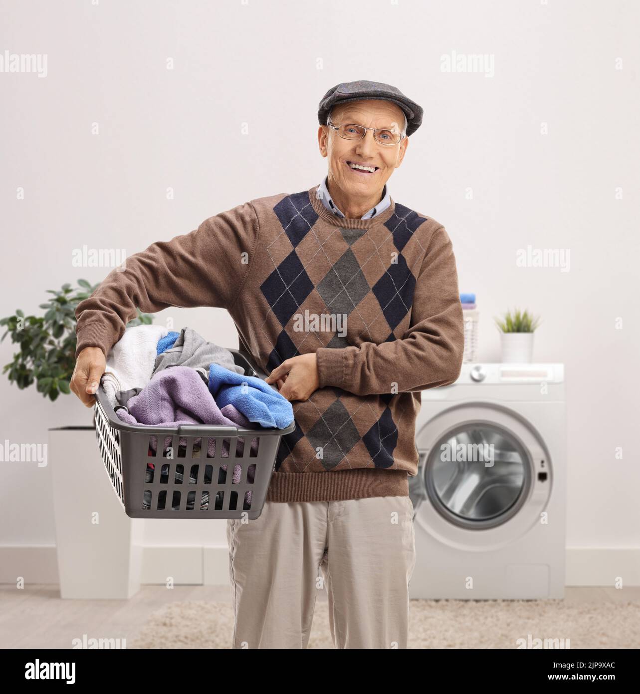 Uomo anziano sorridente che tiene un cesto di biancheria pieno di vestiti all'interno di un bagno Foto Stock