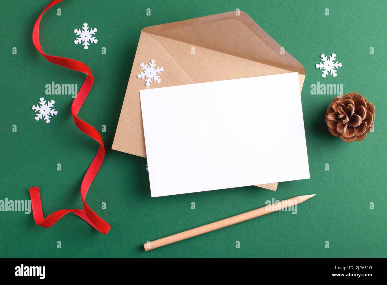 Beffardo di auguri di stagione con cartolina vuota, busta kraft, matita di legno e decorazione di Natale su sfondo verde. Foto Stock