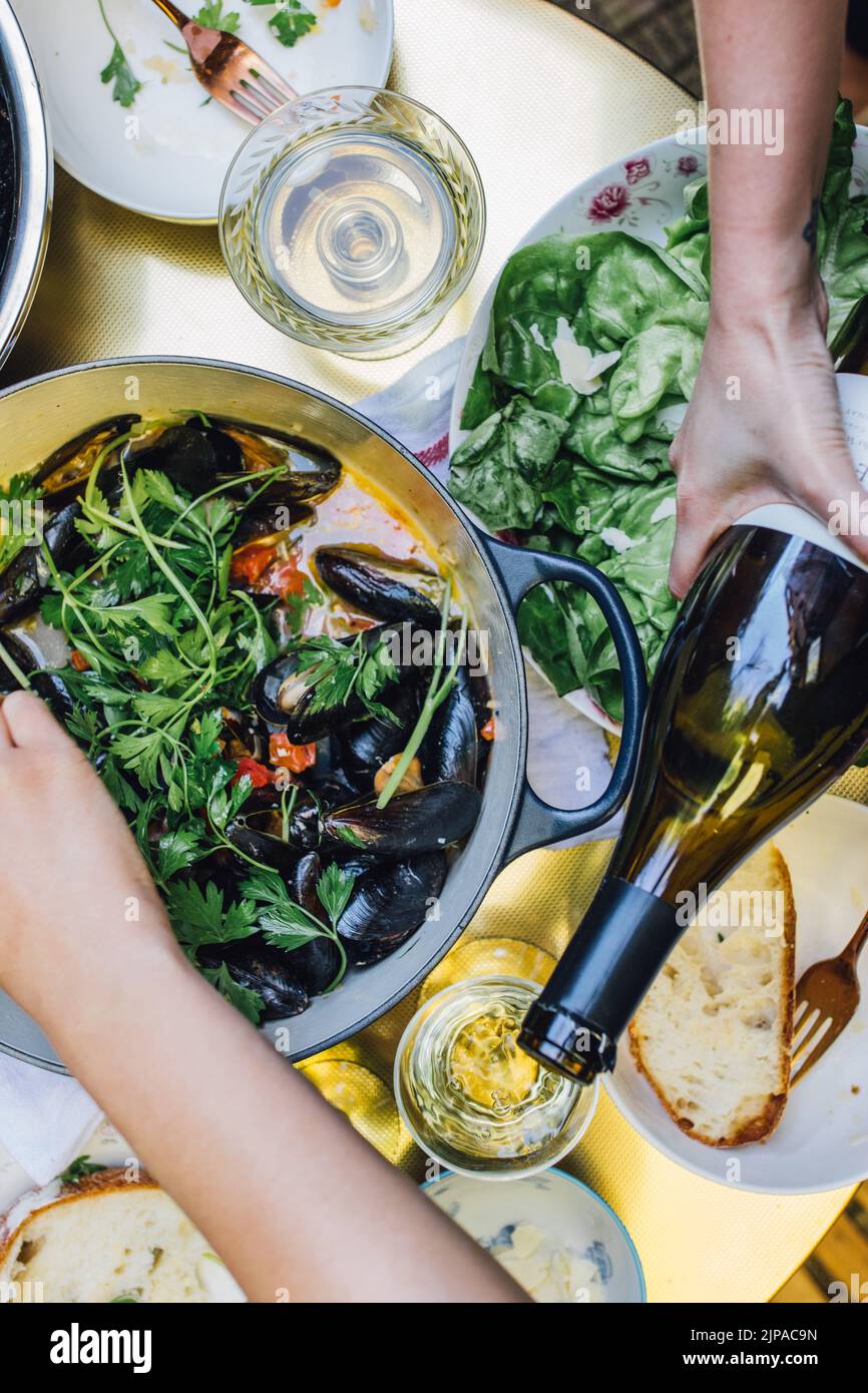 Vista ad alto angolo della cena, vino bianco versato e una pentola di cozze con pomodoro, prezzemolo su tavolo d'oro Foto Stock