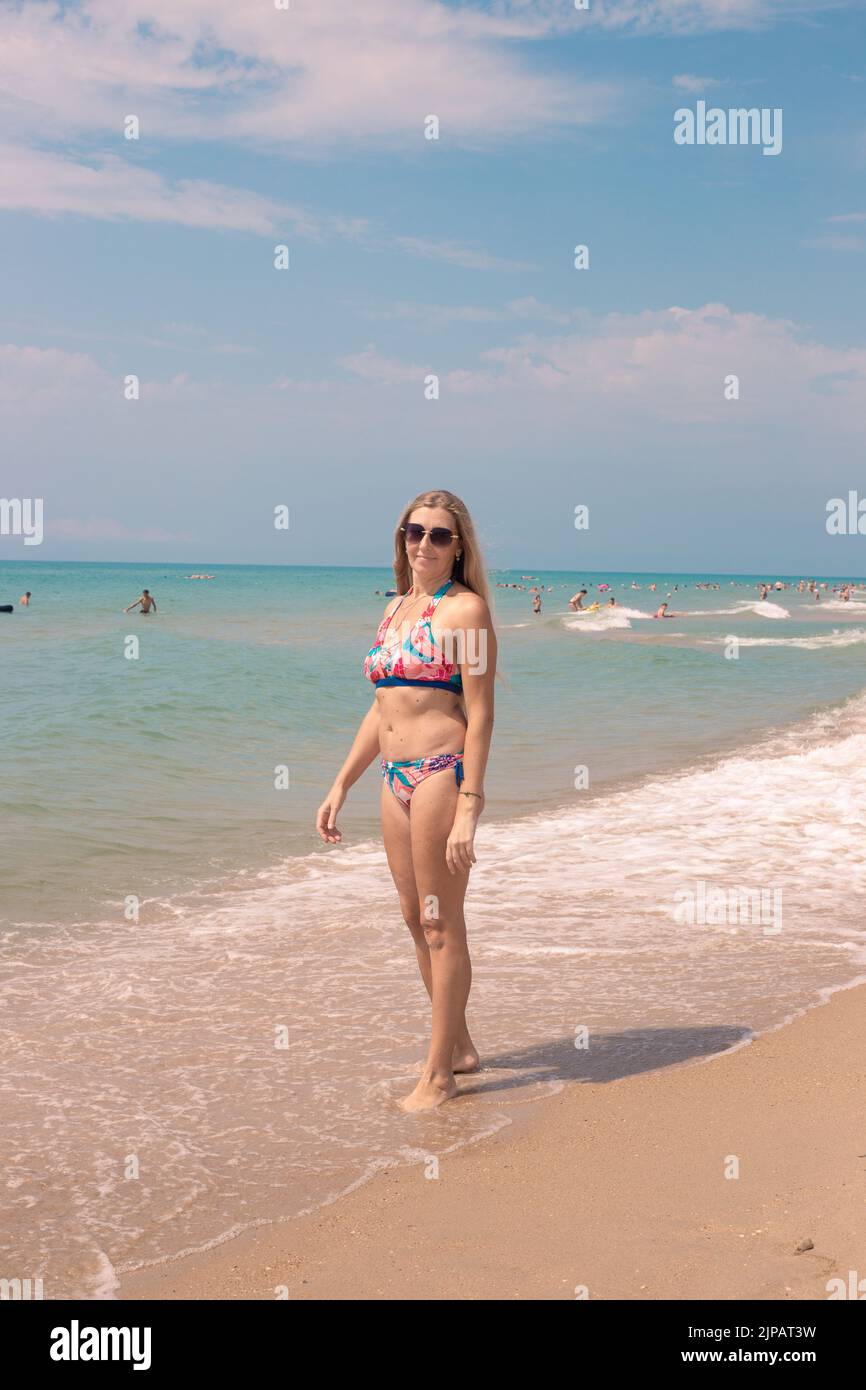 Una donna adulta con lunghi capelli biondi si trova sulla riva del mare blu in una giornata di sole. Viaggi e vacanze estive. Foto Stock