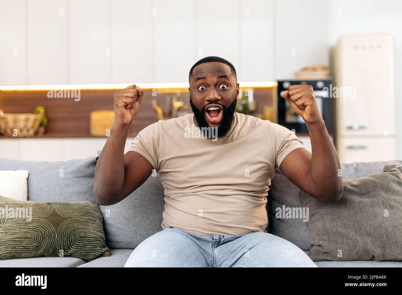 Eccitato gioioso attraente giovane afro-americano, seduto sul divano in soggiorno, eccitato per il successo con le braccia alzate, celebrando la vittoria sorridendo felicemente, urlando. Concetto vincente Foto Stock