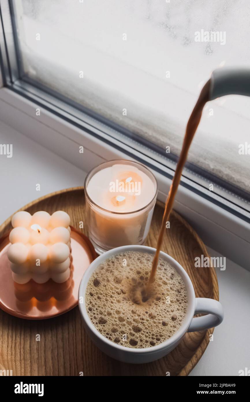 Versare cacao caldo o caffè con latte in una tazzina.Inverno vita morta con bevande calde e candele Foto Stock