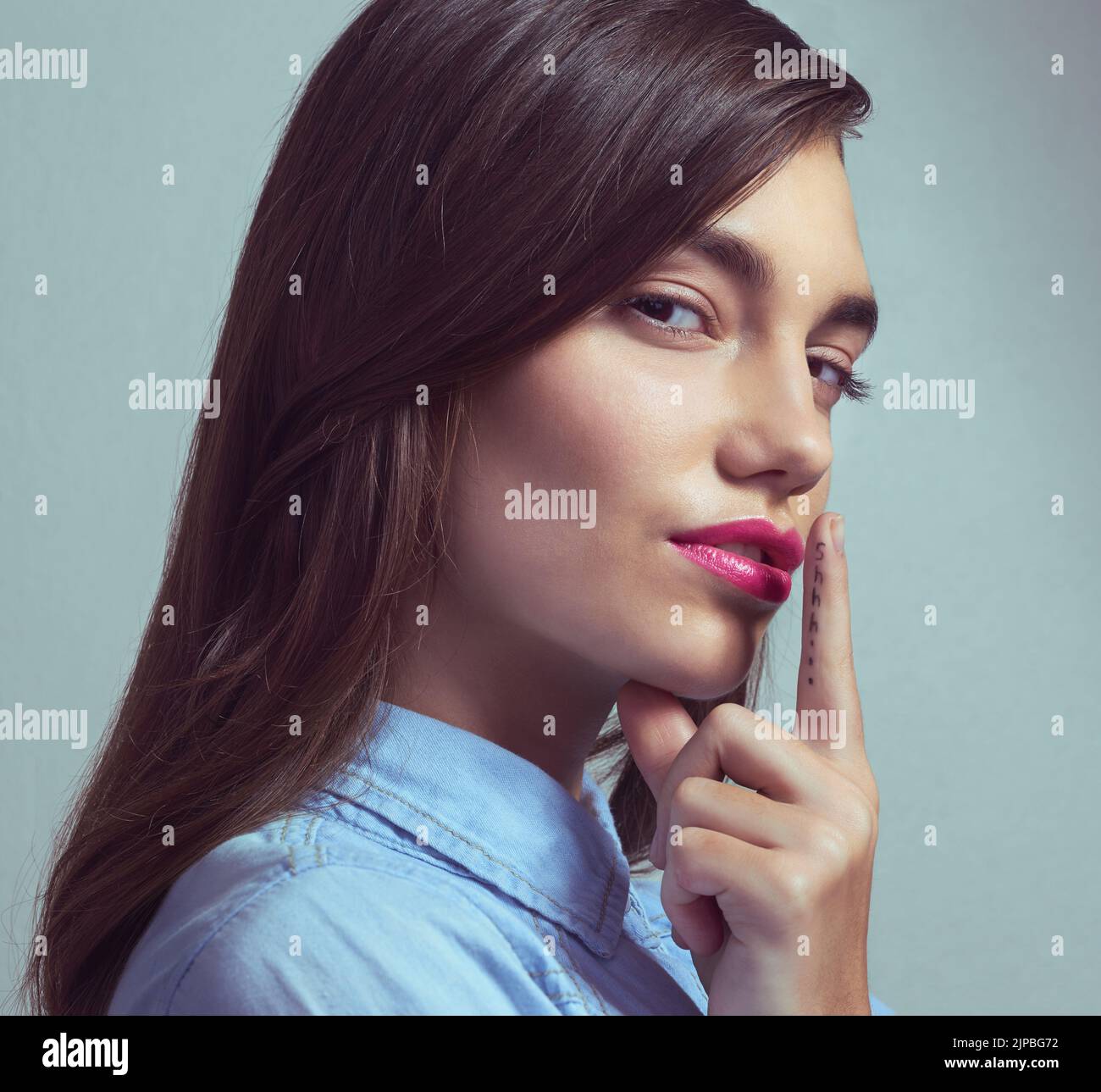 Youre giurato alla segretezza. Studio ritratto di una giovane donna attraente posa con il dito sulle labbra su uno sfondo grigio. Foto Stock
