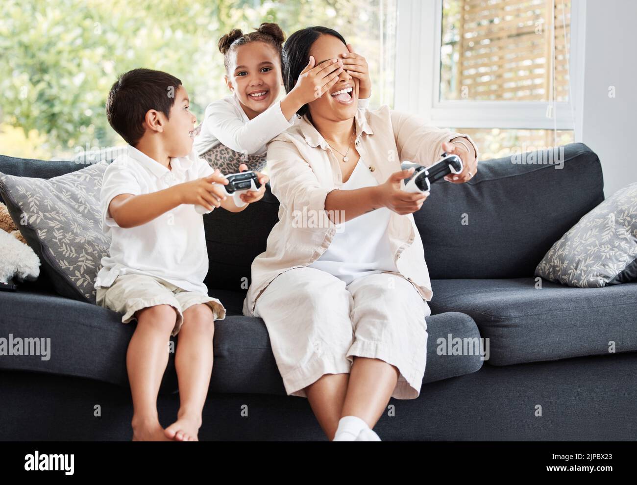 Famiglia, videogiochi e imbrogliare la bambina che copre gli occhi delle madri per permettere al fratello di vincere sul divano a casa. Intrattenimento, joystick e divertimento Foto Stock