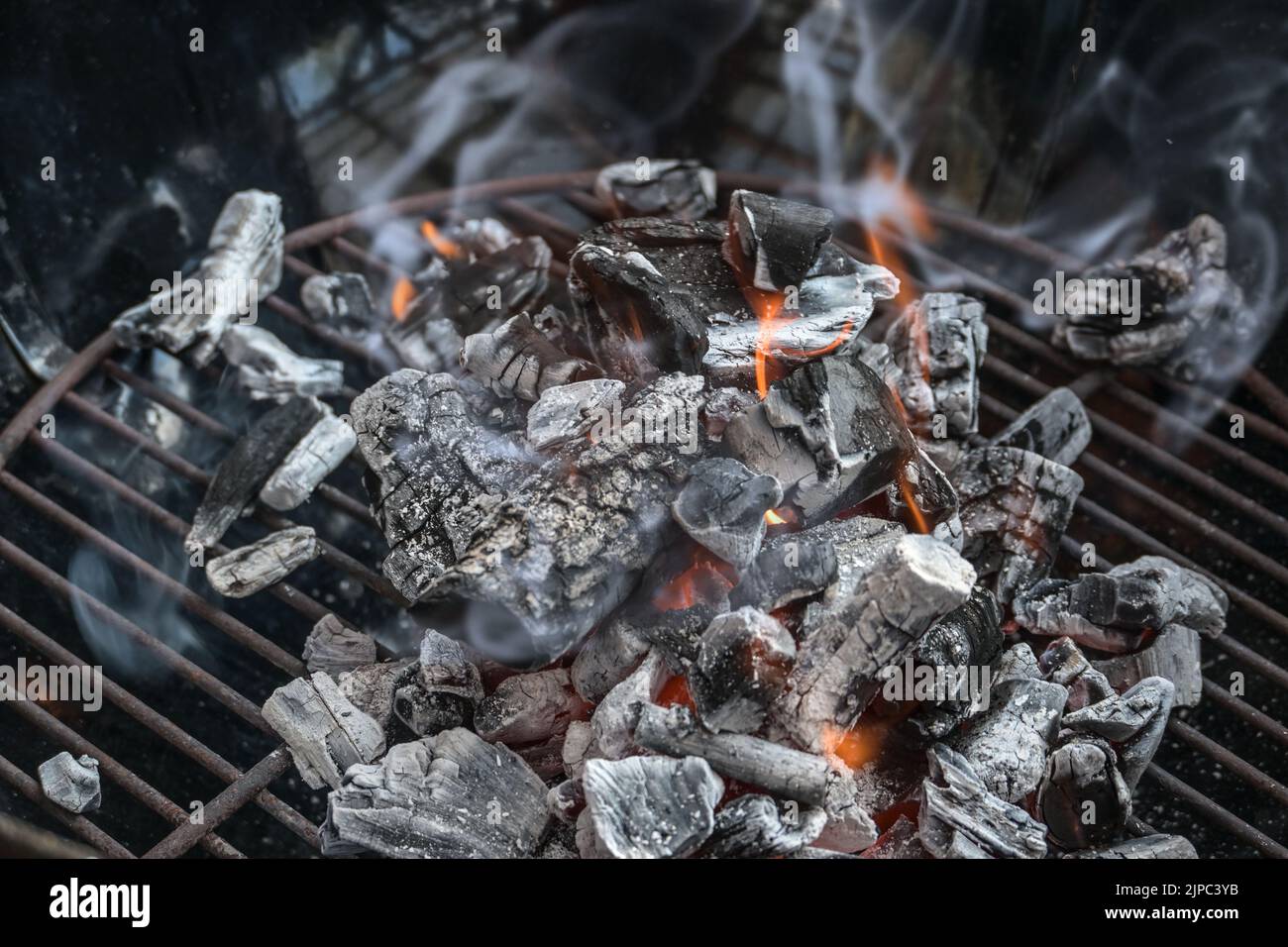 Carbone di legna incandescente con piccole fiamme e fumo in una griglia nera, preparazione per un barbecue, spazio copia, fuoco selezionato, profondità di campo stretta Foto Stock
