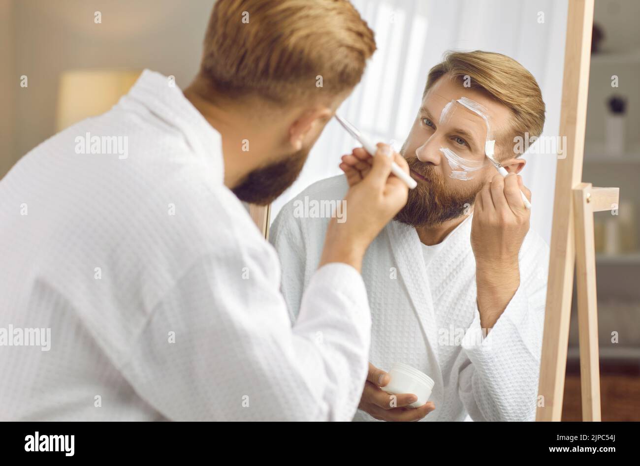 Bell'uomo adulto in accappatoio bianco guardando in specchio e applicando maschera di argilla bianca Foto Stock