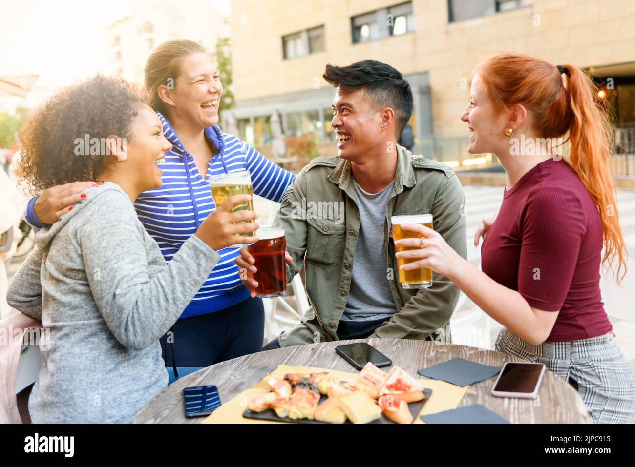 Donna felice ridendo e abbracciando diversi amici con la birra seduto al tavolo da pub con spuntini e smartphone sulla strada della città Foto Stock