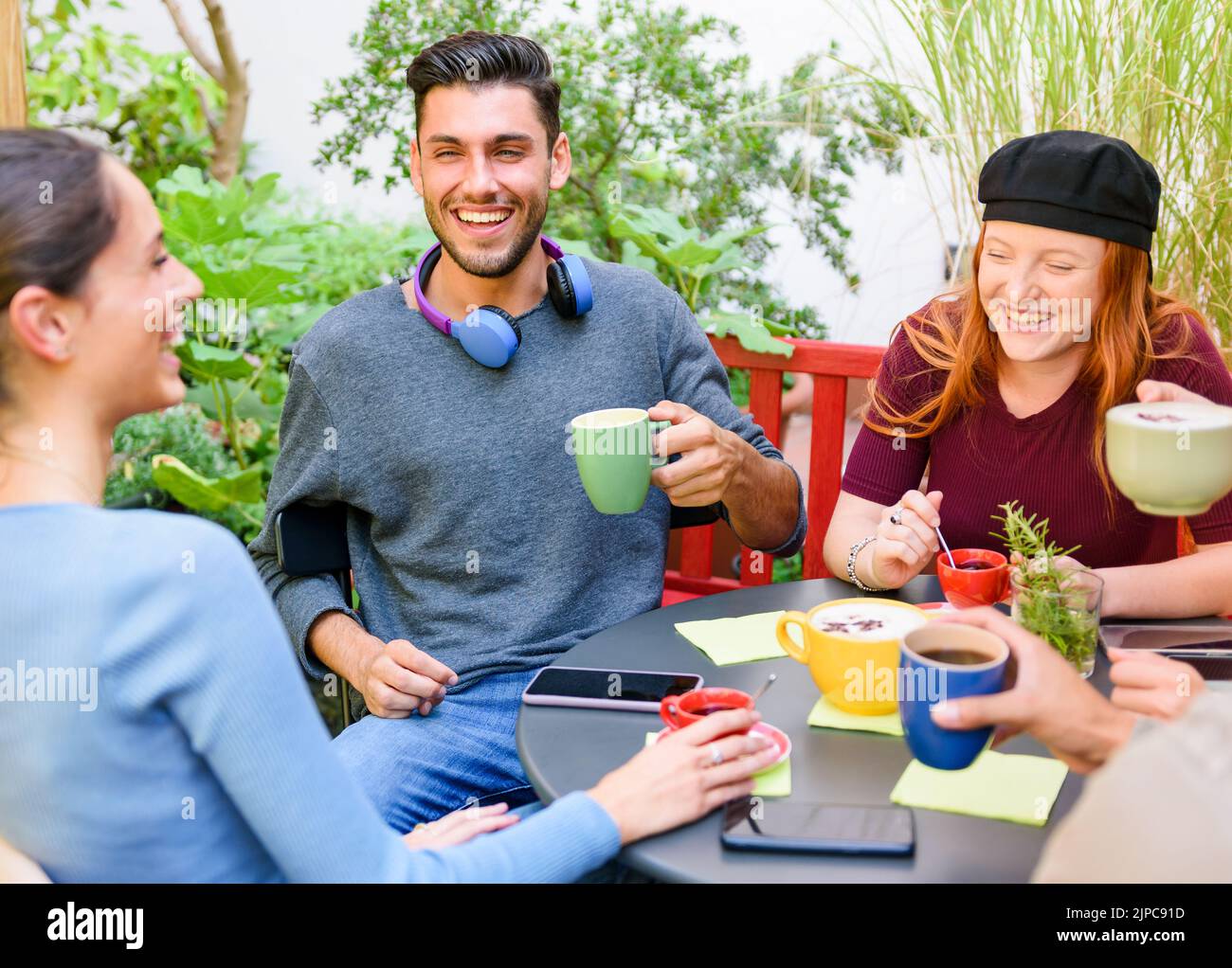Angolo alto di giovani uomini e donne allegri che bevono caffè e ridono di scherzo mentre si siedono intorno al tavolo durante la pausa caffè in giardino Foto Stock