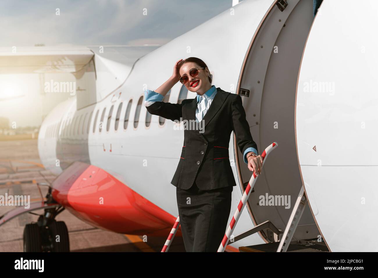 Donna assistente di volo in occhiali da sole in piedi sulle scale dell'aereo all'aeroporto e guardando la macchina fotografica Foto Stock